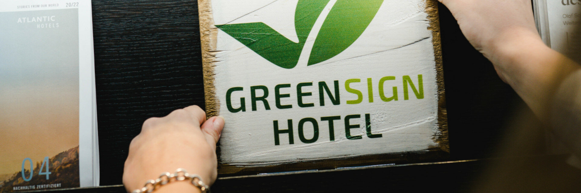 GreenSign Hotel | ATLANTIC Hotel Lübeck is Level 4 certified Person holding a wooden sign with a green leaf and "Greensign Hotel" next to ATLANTIC Hotel brochure.