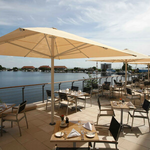 Terrace in the ATLANTIC Hotel Wilhelmshaven Terrace with tables, chairs and parasols, overlooking a peaceful lake and a town in the background.