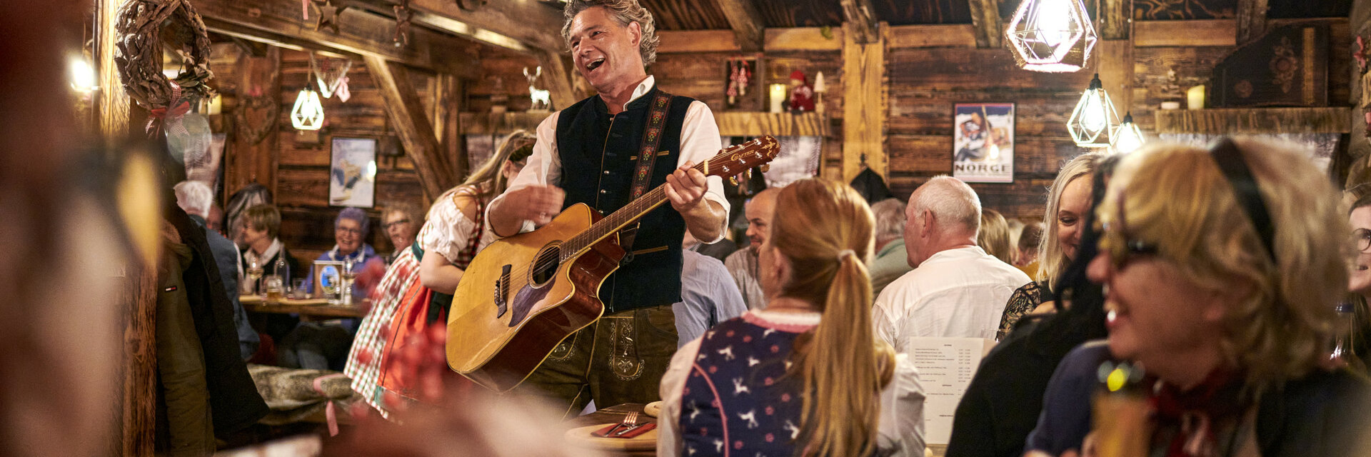 Celebrate in the alpine hut at the ATLANTIC Grand Hotel Bremen Cozy hut with a musician playing the guitar, surrounded by laughing guests in traditional dress.