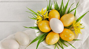 Weiße Schale mit gelben und weißen Ostereiern, dekoriert mit gelbem Blütenschmuck und grünen Blättern auf weißem Tuch.