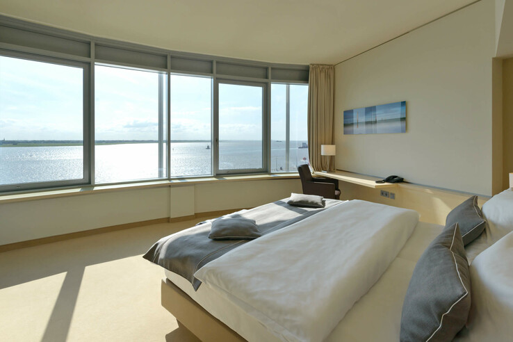 Interior view of the Suite in the ATLANTIC Hotel SAIL City in Bremerhaven with an amazing view over the Weser Bright hotel room with large bed and panoramic window overlooking the water.
