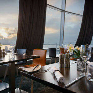 Gedeckte Tische im Restaurant STROM des ATLANTIC Hotel SAIL City Bremerhaven Modernes Restaurant mit gedeckten Tischen, großen Fenstern und Blick auf Sonnenuntergang über dem Wasser im ATLANTIC Hotel.