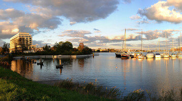 Exterior view ATLANTIC Hotel Wilhelmshaven Hotel on the water with sailing boats in the harbor, surrounded by trees, under a cloudy sky at sunset.