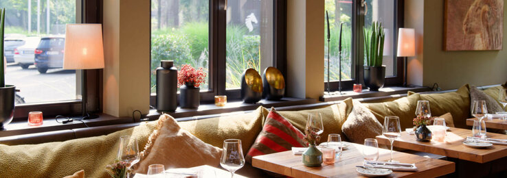 Cozy restaurant in the ATLANTIC Hotel Landgut Horn with covered wooden tables, cushions and a view of the green outdoor area.