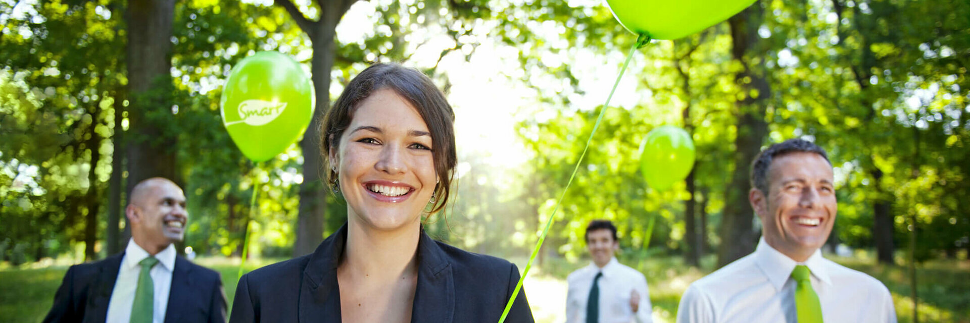 Tagen in den ATLANTIC Hotels Lächelnde Menschen in Anzügen halten grüne Ballons im sonnigen Wald.