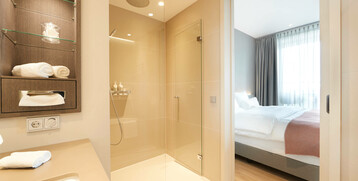 Bathroom of the Balcony Suite at the Atlantic Grand Hotel Bremen Modern hotel bathroom with shower, washbasin and view into the bright bedroom with double bed at the ATLANTIC Grand Hotel Bremen.