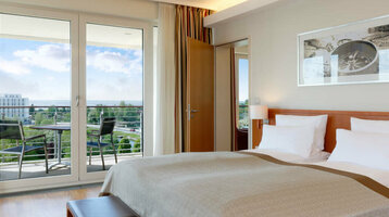 Übernachtungsangebot 5 für 4 Helles Hotelzimmer mit Doppelbett, Holzboden, TV, Balkon mit Stühlen und Blick auf grüne Landschaft.