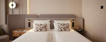 Modern hotel room with two single beds placed side by side, light-coloured duvets and decorative cushions with floral patterns. On the left is a small wooden table with magazines, on the right a round side table with a drink and remote control. Subtle wall lighting and warm, neutral colours characterise the room.