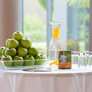 ATLANTIC Hotel Universum SMART Gesundheit Ein Tisch mit grünen Äpfeln, Wasserkaraffe mit Orangen, Gläsern und Broschüren in einem hellen Raum.