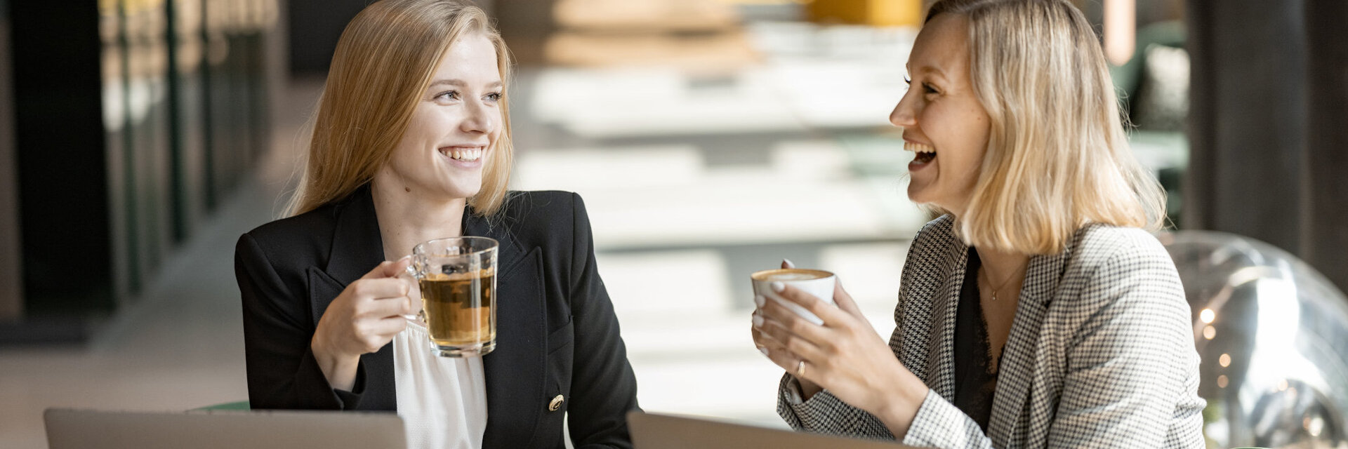 Zwei Frauen lachen und sitzen vor Laptops während sie Kaffee in der Hand halten.