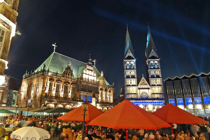 Musikfest Bremen Lively square in front of an illuminated historic building and church at night, with red parasols and crowds of people.