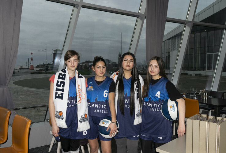 Co-operation girls' team Eisbären Bremerhaven U14 with ATLANTIC Hotel Sail City Four basketball players in ATLANTIC Hotels jerseys pose in front of a window with a view of the harbor.