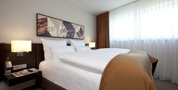 Double bed in the Studio of the ATLANTIC Grand Hotel in Bremen Modern hotel room with double bed, white bed linen, brown bedspread, artwork on the wall and natural light through the window.