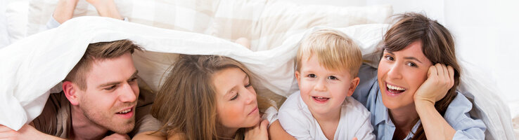 Family offer ATLANTIC Hotel Universum Family smiling happily under a comforter in a bright hotel room at the ATLANTIC Hotel Universum in Bremen.