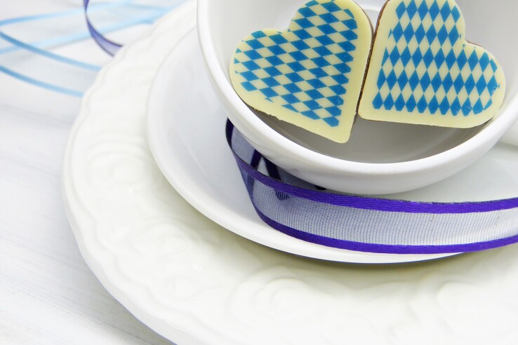 Munich Heart White cup with two heart-shaped cookies, blue and white checkered, decorated with a purple ribbon on a saucer.