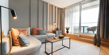 Balcony Suite living area at the Atlantic Grand Hotel Bremen Modern hotel room with gray sofa, colorful cushions, coffee table, lamp and balcony view through large windows.