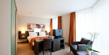 Interior view of the seating area in the Executive Room of the ATLANTIC Grand Hotel in Bremen Bright hotel room with double bed, desk, TV, armchairs and large windows with orange curtains.