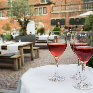 Enjoy cool drinks in the Hofgarten of the ATLANTIC Grand Hotel Bremen Three glasses of red wine on a tray in the outdoor area of the ATLANTIC Grand Hotel Bremen, with tables in the background.