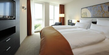 Sleeping area Suite in the ATLANTIC Grand Hotel Bremen Modern hotel room with large bed, art on the wall, TV, armchair and floor-to-ceiling windows with curtains.