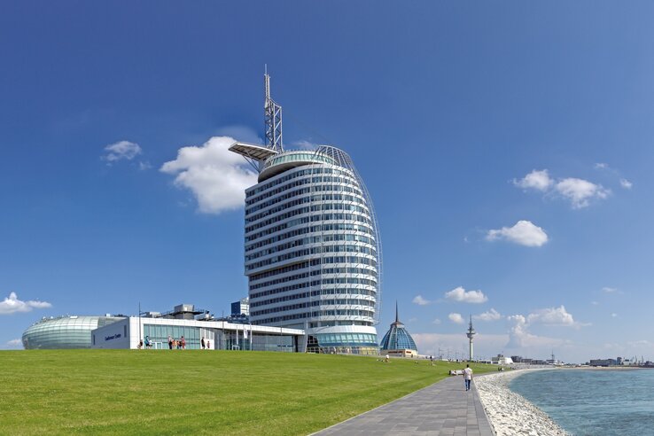 Außenansicht auf das Gebäude des ATLANTIC Hotel SAIL City von der Küste aus Modernes Hotelgebäude am Meer mit geschwungener Fassade, umgeben von Rasenflächen und einem Gehweg entlang der Küste.