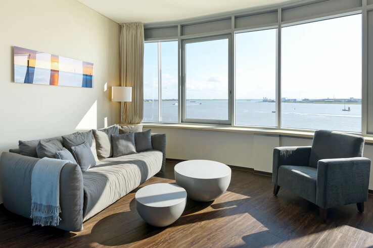 Interior view of the Suite in the ATLANTIC Hotel SAIL City in Bremerhaven with an amazing view over the Weser Bright hotel room with gray sofa, armchairs, round tables and panoramic window overlooking the water.