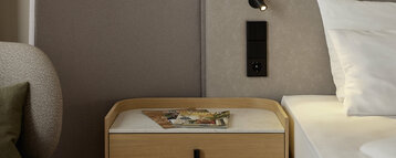 Close-up of a modern hotel room: next to the bed is a small wooden bedside table with a light-coloured top, on which lies a magazine. On the upholstered wall behind the bed is a small reading lamp and a black control panel.